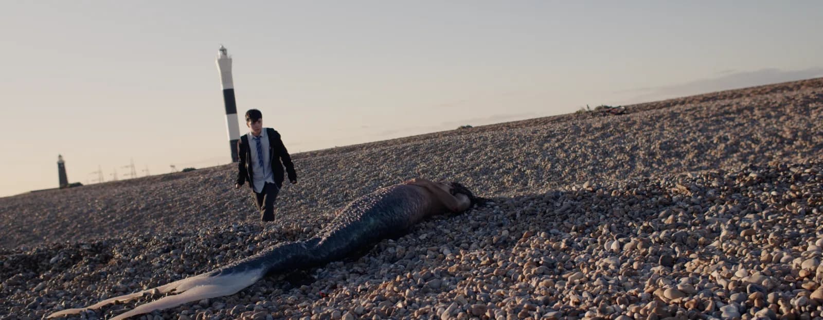A boy in a school uniform walks across a shingle beach towards a mermaid lying on the stones, with a black and white lighthouse behind him